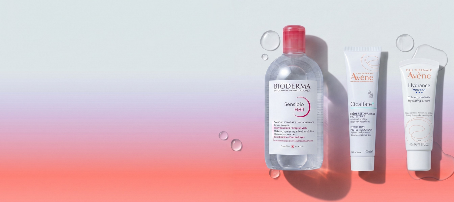Three skincare products arranged on a clean studio background: a clear Bioderma Sensitivity Red cleansing bottle with a pink cap, a white Aveeno Crèche tube, and a white Aveeno Hydroccra tube, with floating bubble graphics and a soft pink-to-white gradient background.